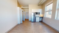 Located at 61 E 21st St in Chicago's vibrant Near South Side neighborhood, this thoughtfully designed single-floor unit combines modern finishes with functional living. The open-concept main living area is bathed in natural light from a row of tall windows that frame views of the surrounding brick exterior, adding an urban industrial character to the warm, neutral interior.

The compact yet well-appointed kitchen is fitted with stainless steel appliances, including a full-size refrigerator, built-in microwave, dishwasher, and a cooktop with sink, all complemented by crisp white cabinetry that extends to the ceiling for generous storage. Exposed ductwork overhead adds an industrial loft aesthetic that contrasts pleasantly with the clean, light-toned walls.

Wood-style luxury vinyl flooring runs seamlessly throughout the unit, lending warmth and continuity to every room. The bedroom features a spacious walk-in closet with built-in shelving and rod storage, providing ample organization. A hallway leads to the full bathroom, which includes a soaking tub and shower combination, a wall-mounted vanity with sink, and a mirrored medicine cabinet. The elevated ceilings throughout the unit enhance the overall sense of openness and airiness.