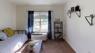 This single-floor unit offers a thoughtfully organized layout that makes efficient use of its footprint. The main living and sleeping area is defined by a low half-wall partition, which provides a subtle sense of separation while maintaining an open feel throughout the space. Soft carpeting underfoot adds warmth and comfort, while a large window dressed with navy curtains and horizontal blinds allows natural light to fill the room and offers a view of the surrounding exterior. A wall-mounted HVAC unit ensures year-round climate control.

The compact kitchen is clean and functional, featuring white cabinetry, a coil-top electric range, an over-the-range microwave, and a stainless steel sink with ample counter space — everything needed for everyday meal preparation. A small breakfast bar area with a stool connects the kitchen to the living space, enhancing the unit's versatility.

The bathroom is accessible via a warm-toned wooden door and is finished with light tile flooring, a wall-mounted sink, and a toilet. Framed botanical artwork and a mirror contribute to a calm, tidy atmosphere. The overall palette of the unit is neutral and bright, with white walls throughout, creating a clean and welcoming environment well-suited for comfortable everyday living.