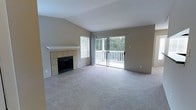 This single-floor residence offers a welcoming living area defined by vaulted ceilings that lend a sense of openness and airiness to the space. The room is finished with neutral-toned wall-to-wall carpeting, providing a comfortable and cohesive foundation throughout.

A standout feature of the living room is the fireplace, framed by a clean tile surround and hearth in warm earth tones, topped with a simple white mantel. The design is understated yet functional, serving as a natural focal point within the room.

Natural light is a prominent characteristic of this space, drawn in through a large window fitted with horizontal blinds and a sliding glass door that together illuminate the interior with abundant daylight. The sliding door opens directly onto a private balcony enclosed by a classic white railing, overlooking lush, mature tree coverage that provides a serene, nature-filled backdrop. The seamless connection between the interior living area and the outdoor balcony enhances both the sense of scale and the overall ambiance of the home, making it feel open and connected to its natural surroundings.