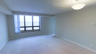 This single-floor unit presents a clean, open living space finished with plush wall-to-wall carpeting in a light neutral tone. The room is characterized by its generous proportions, with smooth, light-colored walls and white baseboards lending a crisp, uniform aesthetic throughout. A large, multi-pane picture window dominates one wall, drawing in substantial natural light and framing a notable elevated view of the surrounding urban cityscape below — suggesting a high-floor position within a larger building. The window's wide span ensures the room remains well-lit during daylight hours, contributing to an airy and open ambiance. Ceiling-mounted flush lighting provides additional illumination, and the textured ceiling adds a subtle architectural detail. The neutral palette of soft grey-green walls paired with the light carpet creates a calm and adaptable backdrop suitable for a variety of interior styles. Overall, the space presents a straightforward, functional layout with a standout feature in its elevated city views, offering a quiet and understated environment.