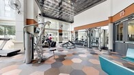 This 1,130-square-foot fitness center is a single-floor facility designed to accommodate a comprehensive workout experience. The space is defined by its dramatic double-height ceilings adorned with a decorative embossed tin ceiling treatment, lending an upscale architectural character to the room. Floor-to-ceiling windows line multiple walls, flooding the interior with abundant natural light and offering open views of the surrounding urban streetscape and greenery.

The flooring features a bold, eye-catching pattern of large hexagonal tiles in muted tones of blush, gray, and taupe, transitioning to rubberized mats in the dedicated free weights and strength training areas. The cardio zone is equipped with treadmills, elliptical machines, rowing machines, and stationary bikes. The strength training section includes a multi-station cable machine, a full dumbbell rack, and adjustable weight benches. Stability balls are neatly stored on a dedicated rack near the windows.

Warm terracotta accent bands run along the upper walls, complementing the neutral color palette and adding visual depth. Wall-mounted fans and flat-screen televisions are positioned throughout for comfort and entertainment. A seating area near the entrance provides a welcoming lounge space, complete with contemporary chairs. Large glass entry doors complete this bright, modern, and functional fitness facility.