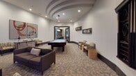 Located at 300 Blue Moon Xing in Pooler, Georgia, this single-floor community amenity space is thoughtfully designed to serve as a social and recreational hub. The layout seamlessly blends a game room and lounge area into one cohesive environment, anchored by a full-size billiards table positioned beneath a sleek pendant light fixture. An architecturally distinctive curved, layered ceiling detail adds visual depth and character to the billiards area, while recessed lighting throughout ensures the space is well-lit. The lounge zone features a comfortable arrangement of striped accent chairs, a dark sofa, and a coffee table set atop patterned carpeting, creating an inviting area for relaxing or socializing. A wall-mounted flat-screen television is positioned on a teal accent wall, complemented by a credenza below for added functionality. A wall-mounted cue rack keeps billiards equipment neatly organized. Contemporary artwork and decorative accents are displayed throughout, contributing to the polished, modern aesthetic. Dark-framed doorways connect the space to adjoining areas, including a fitness center visible through one of the passages, offering a glimpse of exercise equipment beyond. The overall ambiance is modern and welcoming, suited for both casual gatherings and active recreation.