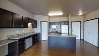 This single-floor space centers around a thoughtfully designed kitchen that serves as the focal point of the layout. The kitchen features a dual-tone cabinetry scheme, combining rich dark-brown lower and upper cabinets along one wall with gray-toned upper cabinets near the cooking area, creating a visually layered aesthetic. Granite-style countertops run throughout, providing ample workspace and a polished finish. Stainless steel appliances, including a refrigerator, electric range with overhead microwave, and dishwasher, equip the kitchen with modern functionality. A peninsula island with matching countertop offers additional prep space and potential for casual dining or serving. Hardwood-style flooring extends across the space, adding warmth and continuity. Recessed and overhead lighting ensures the kitchen remains well-lit. Multiple doorways visible from the kitchen suggest a practical, connected floor plan. The overall ambiance is clean, contemporary, and functional, making the kitchen a versatile hub suited for both everyday cooking and entertaining.