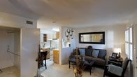 This single-story space in Euless, Texas, offers an open-concept layout that seamlessly connects the living area and kitchen, creating a cohesive and functional flow. The living room is a comfortable and inviting space, highlighted by a large framed mirror that enhances the sense of openness and reflects light throughout the room. Decorative wall accents, including circular metalwork, add a contemporary touch to the neutral-toned walls. The seating area features a gray sofa adorned with blue and gray throw pillows, complemented by a glass-topped coffee table that adds a sleek, modern element. Decorative vases and dried botanicals provide additional styling throughout the space. The kitchen is partially open to the living area, separated by a half-wall that maintains visual connectivity. It features warm wood-toned cabinetry, white appliances including a refrigerator and range, and a range hood above the cooktop. A bar-height seating area along the kitchen partition offers a casual dining option. Light-colored flooring runs throughout both areas, contributing to the bright and airy ambiance of this well-appointed single-floor space.