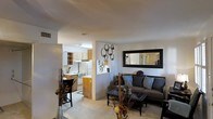 This single-story space in Euless, Texas, offers an open-concept layout that seamlessly connects the living area and kitchen, creating a cohesive and functional flow. The living room is a comfortable and inviting space, highlighted by a large framed mirror that enhances the sense of openness and reflects light throughout the room. Decorative wall accents, including circular metalwork, add a contemporary touch to the neutral-toned walls. The seating area features a gray sofa adorned with blue and gray throw pillows, complemented by a glass-topped coffee table that adds a sleek, modern element. Decorative vases and dried botanicals provide additional styling throughout the space. The kitchen is partially open to the living area, separated by a half-wall that maintains visual connectivity. It features warm wood-toned cabinetry, white appliances including a refrigerator and range, and a range hood above the cooktop. A bar-height seating area along the kitchen partition offers a casual dining option. Light-colored flooring runs throughout both areas, contributing to the bright and airy ambiance of this well-appointed single-floor space.