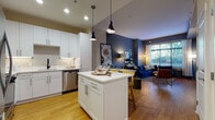 This well-designed single-floor residence offers a cohesive and contemporary living experience through a thoughtfully planned open-concept layout. The kitchen is a standout feature, equipped with crisp white shaker cabinetry, quartz countertops, stainless steel appliances including a gas range, built-in microwave, refrigerator, and dishwasher, all complemented by a matte black faucet and under-cabinet lighting. A central kitchen island with seating provides additional prep space and a casual dining option, anchored by sleek pendant lighting overhead. The kitchen flows seamlessly into the dining and living areas, which are characterized by warm hardwood-style flooring, elevated ceilings, and a bold navy accent wall. Large windows invite abundant natural light into the living room, creating a bright and inviting atmosphere with views of surrounding greenery. The bedroom is a calm retreat finished in soft gray tones, featuring hardwood-style flooring, generous window placement, and ample wall space. The bathroom includes a full soaking tub and shower combination with tile surround. Throughout the residence, a consistent design palette of navy, gray, and white lends the space a modern yet warm aesthetic.