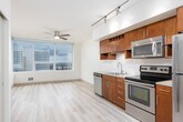 This single-floor unit is designed with a clean, contemporary aesthetic that emphasizes open sightlines and natural light. Light-toned wood-style flooring runs continuously throughout the space, creating a cohesive and airy feel from room to room.

The kitchen serves as a functional centerpiece, featuring rich warm-toned wood cabinetry extending to the upper walls, stainless steel appliances including a refrigerator, range, microwave, and dishwasher, and a tile backsplash. Track lighting overhead ensures strong task illumination. A large window beside the kitchen allows natural light to flow in while offering views of the surrounding cityscape, complemented by a ceiling fan nearby.

The main living area is generously proportioned and open, benefiting from the same natural light and city views visible through floor-to-ceiling windows. The layout flows naturally from the kitchen into this open living space.

A hallway connects the living area to the bedroom and bathroom. The bedroom is a comfortable, well-proportioned room with a sliding closet door and recessed lighting. The bathroom is straightforward and cleanly finished, equipped with a full soaking tub, shower combination with a curved curtain rod, and neutral-toned flooring. Recessed lighting throughout the unit maintains a modern, uncluttered ceiling appearance.