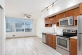 This single-floor unit is designed with a clean, contemporary aesthetic that emphasizes open sightlines and natural light. Light-toned wood-style flooring runs continuously throughout the space, creating a cohesive and airy feel from room to room.

The kitchen serves as a functional centerpiece, featuring rich warm-toned wood cabinetry extending to the upper walls, stainless steel appliances including a refrigerator, range, microwave, and dishwasher, and a tile backsplash. Track lighting overhead ensures strong task illumination. A large window beside the kitchen allows natural light to flow in while offering views of the surrounding cityscape, complemented by a ceiling fan nearby.

The main living area is generously proportioned and open, benefiting from the same natural light and city views visible through floor-to-ceiling windows. The layout flows naturally from the kitchen into this open living space.

A hallway connects the living area to the bedroom and bathroom. The bedroom is a comfortable, well-proportioned room with a sliding closet door and recessed lighting. The bathroom is straightforward and cleanly finished, equipped with a full soaking tub, shower combination with a curved curtain rod, and neutral-toned flooring. Recessed lighting throughout the unit maintains a modern, uncluttered ceiling appearance.