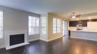 This single-floor space showcases a well-designed open layout that seamlessly connects the living area to the kitchen. The living room is anchored by a clean, white fireplace along one wall, adding a classic yet modern focal point to the space. Dark hardwood floors run throughout, providing a rich contrast against the neutral gray and warm beige wall tones.

Natural light is a defining feature, with multiple windows fitted with white horizontal blinds lining two walls of the living area. A sliding glass door with a grid-pane design allows additional light while providing access to an outdoor area. The open feel is further enhanced by the ceiling fan with integrated lighting, contributing both function and style.

The kitchen, visible from the living area, features dark upper cabinetry paired with a light-colored countertop, creating a balanced contrast. The open sightlines between the living and kitchen spaces make the overall floor plan feel expansive and connected. The combination of modern finishes, ample natural light, and cohesive neutral tones gives this space a clean, contemporary ambiance well-suited for everyday living.