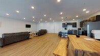 Located in Wisconsin Dells, WI, this single-floor space offers a generous, open-concept layout designed for group gatherings and communal use. The interior is finished with light wood-tone luxury vinyl plank flooring throughout, crisp white walls, and recessed lighting that creates a bright, welcoming ambiance.

The fully equipped kitchen is a standout feature, outfitted with rich dark-stained cabinetry, white quartz countertops, and a full suite of stainless steel appliances including a refrigerator with ice maker, range with microwave, and dishwasher. A central kitchen island with seating provides additional workspace and a casual dining option, while modern pendant lights hang above, adding a contemporary touch.

The open lounge area is generously sized, offering ample room for relaxed seating with multiple gray upholstered sofas and a wall-mounted flat-screen television. The layout allows for flexible arrangement to accommodate groups of varying sizes. A distinctive live-edge wood bar table with matching bench seating adds a rustic, organic contrast to the otherwise modern aesthetic.

A set of interior double doors provides access to an additional area, while an exterior door with an emergency exit sign offers convenient egress. The overall design is clean, functional, and well-suited for events, group retreats, or community gatherings in the heart of Wisconsin Dells.