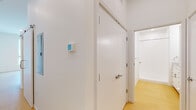 This modern single-floor unit offers a clean, contemporary design throughout, highlighted by warm light hardwood flooring and crisp white walls that create a bright, airy atmosphere. The open-concept layout connects the living area to a well-appointed kitchen, which features white cabinetry, granite countertops, a stainless steel refrigerator, dishwasher, range, and microwave — providing a fully equipped space for everyday living. The living area benefits from large windows that flood the room with natural light while offering views of the surrounding urban landscape, including neighboring buildings and tree canopy. Additional rooms include flexible spaces with clean white finishes and the same consistent hardwood flooring, suitable for use as bedrooms or a home office. A hallway with smart thermostat access connects the rooms efficiently, and a walk-in closet provides practical storage. The bathroom is finished with a granite-topped vanity, white cabinetry with chrome hardware, a large mirror with overhead lighting, and a walk-in shower with white tile surround. Throughout the unit, the design emphasizes simplicity and functionality, with a neutral palette and modern fixtures that create a cohesive, move-in-ready environment.