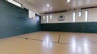 Located at Highland Club Apartments in Watervliet, NY, this dedicated indoor basketball facility offers residents a full-size regulation court designed for serious recreational play. The court features a classic light-colored hardwood-style floor with crisp blue court markings, including a center circle, three-point arcs, free-throw lanes, and key areas. Two regulation backboards and hoops are mounted at either end of the court, providing a complete playing environment.

The space is defined by its impressive ceiling height, which comfortably accommodates full gameplay without restriction. The walls are finished in a bold two-tone scheme — dark forest green on the lower half and a clean off-white above — giving the facility a sharp, athletic aesthetic consistent with the Highland Club branding displayed prominently on the walls. Recessed ceiling lighting ensures even, bright illumination throughout the court, while a series of narrow vertical windows positioned high along the walls allow natural light to supplement the interior lighting.

Access is provided via a dedicated entry door along one end wall. The overall design is functional and purpose-built, making this an excellent amenity for residents seeking an active lifestyle. The facility's scale, quality markings, and dual-hoop setup make it suitable for both casual pickup games and more organized play.