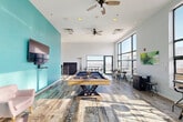 Located at 221 King St in downtown Madison, Wisconsin, this single-floor amenity lounge offers a generously sized, open-plan social space designed for recreation and relaxation. The room is defined by soaring ceilings and floor-to-ceiling windows along the exterior wall, flooding the interior with abundant natural light and framing panoramic views of the Madison skyline and surrounding cityscape. A bold teal accent wall serves as the visual centerpiece of the lounge, anchoring a wall-mounted flat-screen television and a pair of soft pink accent chairs. At the heart of the room sits a substantial wood-framed pool table with slate playing surface, providing a focal point for social gatherings. The distressed, mixed-pattern hardwood flooring adds a rustic-industrial character that complements the modern aesthetic throughout. High-top communal tables with industrial-style stools are arranged near the expansive windows, offering an ideal setting for casual meetings or socializing with a view. Ceiling fans are strategically placed to ensure comfortable airflow across the space. Access to an adjoining outdoor terrace is available through glass doors, further extending the entertainment area. A hallway connects to additional building amenities, completing this well-appointed rooftop gathering space.