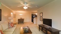 This single-floor residence showcases an open-concept floor plan that seamlessly connects the living, dining, and kitchen areas into one cohesive, flowing space. The living room is anchored by a ceiling fan with a built-in light fixture and features plush wall-to-wall carpeting in a neutral beige tone, contributing to a warm and inviting atmosphere. Framed artwork adorns the walls, adding a decorative touch alongside a media console and flat-screen television.

The dining area sits just beyond the living space, illuminated by a classic chandelier that adds a traditional elegance to the room. A wooden dining table and chairs provide a comfortable setting for meals. The front entry door is visible from this vantage point, contributing to the open and airy feel of the layout.

The kitchen, partially open to the main living area via a breakfast bar, features dark cabinetry contrasted against lighter countertops and walls. A pendant light above the bar area adds functional and ambient illumination. The overall design palette of warm neutrals, dark wood accents, and consistent flooring throughout creates a unified and comfortable living environment.