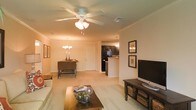 This single-floor residence showcases an open-concept floor plan that seamlessly connects the living, dining, and kitchen areas into one cohesive, flowing space. The living room is anchored by a ceiling fan with a built-in light fixture and features plush wall-to-wall carpeting in a neutral beige tone, contributing to a warm and inviting atmosphere. Framed artwork adorns the walls, adding a decorative touch alongside a media console and flat-screen television.

The dining area sits just beyond the living space, illuminated by a classic chandelier that adds a traditional elegance to the room. A wooden dining table and chairs provide a comfortable setting for meals. The front entry door is visible from this vantage point, contributing to the open and airy feel of the layout.

The kitchen, partially open to the main living area via a breakfast bar, features dark cabinetry contrasted against lighter countertops and walls. A pendant light above the bar area adds functional and ambient illumination. The overall design palette of warm neutrals, dark wood accents, and consistent flooring throughout creates a unified and comfortable living environment.