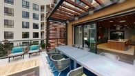 Located at 1055 W Town and Country Rd in Orange, California, this contemporary amenity space seamlessly blends indoor comfort with outdoor living through an open, single-level layout. The interior kitchen area is a standout feature, outfitted with sage-green cabinetry that extends floor to ceiling, dark granite countertops, a deep-set sink, a stainless steel wall oven, and a sleek wood-paneled ceiling with recessed lighting. Terrazzo flooring adds a refined, textural element underfoot, tying together the modern design aesthetic.

Large sliding glass doors connect the interior directly to a covered outdoor terrace, creating a fluid transition between spaces. The terrace is anchored by a generously sized dining table with woven rope chairs, set atop a vibrant blue area rug over light tile. Pendant globe lights hang from a wood-and-steel pergola overhead, providing ambiance during evening gatherings. Additional lounge seating with teal accent cushions and natural wood side tables offer relaxed conversation areas. The terrace overlooks a multi-story residential complex with views of surrounding landscaping, making it a versatile venue for both casual use and organized social events.