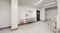 Located at 2 Regal Rd in Toronto's Dovercourt-Wallace Emerson neighbourhood, this shared laundry facility offers a clean, well-organized space designed for practical, everyday use. The facility is composed of a main laundry room and a connecting hallway, together providing a comfortable and functional area for residents.

The main laundry room is bright and efficiently laid out, with white walls and light-toned large-format tile flooring that contribute to a clean, hygienic atmosphere. Fluorescent strip lighting runs across the ceiling, ensuring thorough illumination throughout. A row of modern front-load washing machines lines one wall, complemented by a separate bank of top-load machines along the opposite side, providing ample capacity for multiple users. A stainless steel utility sink is conveniently positioned near the entrance for hand-washing or pre-treatment needs. A payment kiosk and posted usage instructions make operation straightforward for residents.

The adjacent hallway serves as a transitional and utility space, featuring a stainless steel folding/sorting table ideal for managing laundry loads, a bulletin board displaying notices and building information, and a stainless steel waste bin. Two black-framed access doors connect the facility to the rest of the building. The consistent tile flooring and white walls carry through from the main room, maintaining a cohesive and tidy aesthetic throughout the entire facility.
