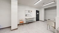 Located at 2 Regal Rd in Toronto's Dovercourt-Wallace Emerson neighbourhood, this shared laundry facility offers a clean, well-organized space designed for practical, everyday use. The facility is composed of a main laundry room and a connecting hallway, together providing a comfortable and functional area for residents.

The main laundry room is bright and efficiently laid out, with white walls and light-toned large-format tile flooring that contribute to a clean, hygienic atmosphere. Fluorescent strip lighting runs across the ceiling, ensuring thorough illumination throughout. A row of modern front-load washing machines lines one wall, complemented by a separate bank of top-load machines along the opposite side, providing ample capacity for multiple users. A stainless steel utility sink is conveniently positioned near the entrance for hand-washing or pre-treatment needs. A payment kiosk and posted usage instructions make operation straightforward for residents.

The adjacent hallway serves as a transitional and utility space, featuring a stainless steel folding/sorting table ideal for managing laundry loads, a bulletin board displaying notices and building information, and a stainless steel waste bin. Two black-framed access doors connect the facility to the rest of the building. The consistent tile flooring and white walls carry through from the main room, maintaining a cohesive and tidy aesthetic throughout the entire facility.