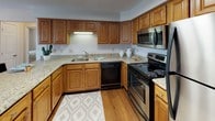 This single-floor residence offers a thoughtfully organized layout that balances comfort and practicality across its living spaces. The living room features neutral-toned carpeting, bright white walls, and ample space for comfortable seating arrangements, with a front entry door and wall-mounted television creating a welcoming, functional environment.

The kitchen is well-equipped with warm oak cabinetry, granite-style countertops, a tile backsplash, and stainless steel appliances including a range and microwave. Hardwood-style flooring runs through this area, adding warmth and easy maintenance. Adjacent to the kitchen, the dining room benefits from hardwood-style flooring, a ceiling fan with light fixture, and a sliding glass door that opens to an outdoor patio, filling the space with natural light.

The bedrooms are carpeted and offer a calm, restful atmosphere. The primary bedroom includes a ceiling fan, ample space, and access to a dedicated bathroom featuring granite-topped oak vanity cabinetry, hardwood-style flooring, and generous counter space. A secondary bedroom also includes closet access and a well-proportioned layout. An additional bathroom mirrors the same granite countertop and oak cabinetry design, maintaining consistency throughout the residence.