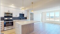 Located at 300 Enterprise Blvd in Bozeman, Montana, this modern 2-bedroom, 2-bathroom unit delivers a well-designed single-level layout with contemporary finishes throughout. The kitchen is a standout feature, showcasing two-tone cabinetry — white upper cabinets paired with light wood lower cabinets — accented by matte black hardware, a dramatic dark marble-style backsplash, quartz countertops, a center island with pendant lighting, and a full suite of stainless steel appliances. Light hardwood flooring flows through the kitchen and living areas, enhancing the bright, open feel of the space. A stacked in-unit washer and dryer adds everyday convenience. The living area benefits from large windows that frame expansive views of the surrounding Montana landscape and distant mountain ranges, flooding the space with natural light. Both bedrooms are carpeted and well-proportioned, with the primary bedroom offering direct access to its own private bathroom. The en-suite bath features large-format tile flooring, a floating wood vanity with quartz countertop, a gold-framed mirror with sconce lighting, and a glass-enclosed walk-in shower. The second bedroom is equally bright, with a window overlooking the open landscape beyond.