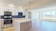 Located at 300 Enterprise Blvd in Bozeman, Montana, this modern 2-bedroom, 2-bathroom unit delivers a well-designed single-level layout with contemporary finishes throughout. The kitchen is a standout feature, showcasing two-tone cabinetry — white upper cabinets paired with light wood lower cabinets — accented by matte black hardware, a dramatic dark marble-style backsplash, quartz countertops, a center island with pendant lighting, and a full suite of stainless steel appliances. Light hardwood flooring flows through the kitchen and living areas, enhancing the bright, open feel of the space. A stacked in-unit washer and dryer adds everyday convenience. The living area benefits from large windows that frame expansive views of the surrounding Montana landscape and distant mountain ranges, flooding the space with natural light. Both bedrooms are carpeted and well-proportioned, with the primary bedroom offering direct access to its own private bathroom. The en-suite bath features large-format tile flooring, a floating wood vanity with quartz countertop, a gold-framed mirror with sconce lighting, and a glass-enclosed walk-in shower. The second bedroom is equally bright, with a window overlooking the open landscape beyond.