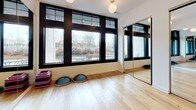 This single-level fitness studio is thoughtfully designed to support a range of movement-based activities. The primary studio room is an open, airy space finished with light natural hardwood flooring that lends warmth and a clean aesthetic throughout. Large, black-framed windows line one wall, flooding the room with natural light and offering views of the surrounding neighborhood. Globe pendant lights suspended from the ceiling provide soft, even ambient illumination to complement the natural light.

Full-length mirrors with sleek black frames are positioned along multiple walls, expanding the visual depth of the room and providing practitioners with clear sightlines for form and technique. The mirrors are partially framed within sliding or fixed panel configurations, adding a polished, studio-quality finish. Fitness props, including stackable step platforms and balance pods, are stored within the studio, indicating its use for group fitness, yoga, or dance instruction.

A set of glass-paneled doors connects the main studio to an adjacent equipment-based gym area, which features rubberized flooring and exercise machines. This seamless transition between zones makes the space highly functional for varied training needs. The overall design is clean, modern, and professional.