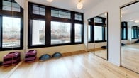 This single-level fitness studio is thoughtfully designed to support a range of movement-based activities. The primary studio room is an open, airy space finished with light natural hardwood flooring that lends warmth and a clean aesthetic throughout. Large, black-framed windows line one wall, flooding the room with natural light and offering views of the surrounding neighborhood. Globe pendant lights suspended from the ceiling provide soft, even ambient illumination to complement the natural light.

Full-length mirrors with sleek black frames are positioned along multiple walls, expanding the visual depth of the room and providing practitioners with clear sightlines for form and technique. The mirrors are partially framed within sliding or fixed panel configurations, adding a polished, studio-quality finish. Fitness props, including stackable step platforms and balance pods, are stored within the studio, indicating its use for group fitness, yoga, or dance instruction.

A set of glass-paneled doors connects the main studio to an adjacent equipment-based gym area, which features rubberized flooring and exercise machines. This seamless transition between zones makes the space highly functional for varied training needs. The overall design is clean, modern, and professional.