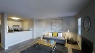 This single-floor space features an open-concept design that connects the living area and kitchen, creating a cohesive and functional layout. The living room is anchored by a neutral-toned sofa accented with yellow and white throw pillows, complemented by a rustic wood-and-metal coffee table atop a dark area rug. A large abstract canvas artwork adorns the grey-painted accent wall, adding a contemporary artistic touch. Warm floor lamps on either side of the seating area enhance the ambient lighting throughout the space. The flooring consists of wood-look planks that extend throughout, providing a clean and modern aesthetic. The textured ceiling adds subtle depth to the interior. Adjacent to the living area, the kitchen features dark countertops with white cabinetry, partially visible through the open layout. The front entryway includes a white paneled door with visible hardware, seamlessly integrated into the living space. Overall, the unit presents a well-coordinated, move-in-ready interior with a contemporary design sensibility.