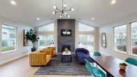 This single-floor space is defined by its striking architectural design, centered around a dramatic vaulted ceiling that peaks above the open-plan living and dining area. The focal point of the main living space is a floor-to-ceiling stacked stone fireplace, clad in textured gray tile, with a wall-mounted television above. A contemporary multi-arm globe chandelier hangs overhead, complementing recessed lighting throughout.

Natural light floods the interior through large sliding glass doors on both sides of the fireplace, offering views of and direct access to outdoor areas. Clerestory windows above the doors further enhance the bright, airy atmosphere. The living area features warm hardwood-style flooring, a patterned area rug, and a mix of bold seating including mustard yellow velvet chairs and a deep navy chaise lounge.

The dining area connects seamlessly to the kitchen, which is accessible via a pass-through counter. The kitchen is fitted with bold navy cabinetry, white countertops, and stainless steel appliances, including a refrigerator. A built-in banquette with matching navy detailing frames a round wood dining table surrounded by teal chairs, creating a cohesive, design-forward aesthetic throughout the space.