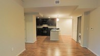 This single-floor space showcases an open-concept layout that integrates the kitchen and living areas into one cohesive environment. The kitchen is a standout feature, appointed with dark espresso cabinetry that extends to the upper and lower levels, providing ample storage. Black appliances, including a range and built-in microwave, complement the cabinetry for a sleek, uniform aesthetic. A peninsula island with a contrasting light-colored countertop serves as both a functional workspace and a natural divider between the kitchen and the adjoining living area. A pendant light fixture hangs above the peninsula, adding a focused decorative element. Recessed lighting throughout the kitchen ensures the space is well-illuminated. The living area benefits from warm hardwood-style flooring that extends throughout, creating visual continuity across the open floor plan. Neutral-toned walls and white baseboards keep the space bright and versatile. A bifold closet door is visible along one wall, offering practical storage solutions. The overall ambiance is modern and functional, with a clean design aesthetic.