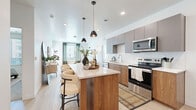 Located at 13500 Antioch Rd in Overland Park, KS, this thoughtfully designed 2-bedroom, 2-bathroom residence offers a cohesive, modern aesthetic throughout its single-floor layout. The open-concept kitchen and living area serves as the heart of the home, anchored by a striking wood-paneled island with quartz countertops and seating for multiple guests. The kitchen is equipped with stainless steel appliances, two-tone cabinetry in warm wood and matte gray, a subway tile backsplash, and pendant lighting that adds a contemporary touch. Light wood-style flooring flows seamlessly through the main living area, which is brightened by large windows dressed with sage green curtains, allowing ample natural light. The primary bedroom is a comfortable retreat featuring plush carpet, a generous walk-in closet with wire shelving, and a large window with exterior views. The second bedroom offers similar warmth and natural light. Both bathrooms are finished with wood-toned vanities, white countertops, and clean tile work. A dedicated laundry room houses a full-size Whirlpool washer and dryer. The overall ambiance is clean, bright, and contemporary, with warm natural materials balanced against a crisp white palette throughout.