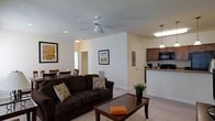 This single-floor residence features an open-concept design that seamlessly integrates the living, dining, and kitchen areas into one functional and inviting space. The living room is anchored by a plush, dark brown sofa accented with decorative throw pillows, positioned around a classic wooden coffee table atop neutral wall-to-wall carpeting. A ceiling fan with a built-in light fixture is centrally mounted, enhancing air circulation and providing ambient lighting. Recessed lighting complements the overall illumination throughout the space. Neutral-toned walls adorned with framed artwork contribute to a clean, understated aesthetic. The dining area, partially visible toward the rear, accommodates a wooden table and chairs, making it well-suited for everyday meals. The kitchen features dark wood cabinetry, dark appliances including a microwave and refrigerator, and countertops with a granite-like finish. Pendant lighting above the kitchen counter adds a polished touch. A door near the kitchen hallway suggests additional rooms beyond the main living area. Crown molding along the upper walls adds a refined architectural detail throughout the open layout.