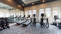This contemporary fitness center is thoughtfully designed to support a full range of workout routines in a single open-plan space. The room features elevated ceilings with a dark recessed ceiling detail and warm wood-paneled accents that contrast elegantly with the neutral walls, creating a polished yet energizing atmosphere. Large windows fitted with roller shades line the perimeter, flooding the space with natural light and offering views of the surrounding neighborhood.

The cardio zone is well-stocked with multiple treadmills, elliptical trainers, and stationary bikes, all equipped with digital displays. A full-length mirror along one wall visually expands the space and is practical for form-checking during workouts. The free weights area features a comprehensive dumbbell rack spanning a wide range of weights, alongside a Matrix power rack with a bench for barbell training. Supplementary equipment includes kettlebells, medicine balls, and TRX suspension trainers, catering to functional and strength training needs alike.

A dedicated amenity station includes built-in wooden shelving, a wall-mounted flat-screen television, a mini refrigerator, and cubby storage for personal items and towels. The patterned carpet tile flooring throughout adds texture and comfort underfoot. A ceiling fan and recessed lighting ensure the space remains comfortable and well-lit at all times, making this fitness center a comprehensive and inviting facility.