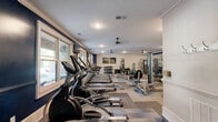 Located at 1000 Flycatcher Way in Arden, NC, this fitness center offers a generously sized, single-floor exercise space designed to support a full range of workout routines. The room is thoughtfully laid out, with distinct zones for cardio, free weights, and strength training.

Along one wall, large windows allow abundant natural light to fill the space while offering views of the surrounding greenery, creating an inviting and energizing atmosphere. A row of treadmills and elliptical machines lines the windowed side, making cardio sessions bright and engaging. Ceiling fans are positioned throughout to maintain comfortable airflow.

The strength training area features a multi-station cable and weight machine setup, complemented by a full Smith machine with plate-loaded barbells. A dedicated free weights section includes a neatly organized dumbbell rack alongside a flat bench, accommodating a variety of resistance exercises. Full-length mirrors on the bold navy accent wall enhance the sense of space and allow for proper form checks during workouts.

The flooring consists of patterned modular carpet tiles in neutral tones, adding both comfort underfoot and a polished aesthetic. Crown molding and classic panel doors lend a refined architectural character that sets this fitness center apart. The space is accessible via multiple entry points and is finished with recessed lighting throughout.