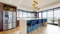 Located at 4757 Hiawatha Ave in Minneapolis, MN, this thoughtfully designed single-floor space delivers a seamless blend of contemporary style and functional living. The open-plan layout connects a well-appointed kitchen and a comfortable living area, all unified by warm light wood flooring throughout.

The kitchen is a standout feature, showcasing rich dark wood cabinetry, a generous white quartz island with navy blue velvet barstools accented in brushed gold, stainless steel appliances, and a striking mid-century modern brass chandelier overhead. Floor-to-ceiling windows flood the kitchen with natural light and frame sweeping outdoor views.

The living area is anchored by a dramatic blue marble-effect accent wall, which frames a mounted flat-screen television and built-in display shelving. Elegant furnishings in neutral tones are punctuated by teal and gold accent pieces, creating a curated, modern aesthetic. Another sculptural brass chandelier mirrors the one in the kitchen, tying the spaces together cohesively.

Large sliding glass doors open directly onto a furnished outdoor terrace, offering expansive views of the Minneapolis skyline and surrounding greenery — a rare and compelling feature that dramatically extends the living space outdoors. Tall narrow windows along the walls further enhance the sense of openness and connection to the exterior environment.