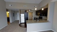 This single-floor residence showcases a thoughtfully designed open-concept layout where the kitchen flows naturally into the living space. The kitchen is a standout feature, equipped with dark espresso cabinetry that contrasts elegantly against the speckled granite countertops. A large peninsula with an undermount sink serves as a functional centerpiece, offering additional workspace and casual seating potential. Stainless steel appliances, including a full-size refrigerator, electric range, and built-in microwave, provide a modern, cohesive aesthetic. Pendant lighting above the peninsula adds warm, focused illumination, complementing the track lighting overhead. The kitchen transitions seamlessly into the carpeted living area, which benefits from the elevated ceiling height that enhances the overall sense of openness. Dark hardwood flooring defines the kitchen zone, providing a clear and stylish distinction between spaces. A hallway visible in the background leads to additional rooms, suggesting a practical and well-organized floor plan. The neutral wall tones throughout create a versatile backdrop suitable for a variety of interior styles.