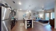 This single-floor residence offers a thoughtfully designed open-concept layout that seamlessly connects the kitchen, dining, and living areas. The kitchen is a standout feature, showcasing dark espresso cabinetry, granite countertops, a mosaic tile backsplash, and a full suite of stainless steel appliances including a refrigerator, range, and microwave. A large central island with bar seating serves as the heart of the space, illuminated by a trio of elegant pendant lights overhead. Rich hardwood-style flooring flows throughout the main living areas, complementing the neutral gray walls and creating a warm, cohesive aesthetic. The living room offers a comfortable, open atmosphere with a ceiling fan, natural light from a large window, and direct access to the entryway. The primary bedroom is generously sized with plush carpeting, natural light from a window, and a ceiling fan. Its en-suite bathroom features a dual-sink granite vanity with dark cabinetry, a glass-enclosed walk-in shower with tile surround, and a spacious walk-in closet. The second bedroom mirrors a similar layout with soft carpeting and natural light. A second full bathroom includes a dual-sink granite vanity. A dedicated laundry area and ample storage complete this well-rounded residence.