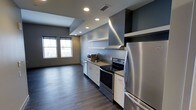 This single-floor space showcases a clean, modern aesthetic with an open-concept design that seamlessly connects the kitchen and living areas. The kitchen is well-appointed with stainless steel appliances, including a refrigerator, electric range, dishwasher, and microwave. White cabinetry provides ample storage, while a bold geometric range hood serves as a striking focal point above the cooktop. Floating shelves add both functionality and visual interest to the kitchen wall. The space features dark hardwood-style flooring that contrasts beautifully against the cool gray and warm beige wall tones. Recessed ceiling lighting is strategically placed throughout, ensuring even illumination across the open floor plan. A raised ceiling detail above the kitchen area adds architectural dimension, enhancing the sense of volume within the space. Two windows in the adjacent living area allow natural light to filter in, creating a bright and welcoming atmosphere. The overall design conveys a contemporary, urban sensibility, balancing practical functionality with modern style.