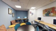 Located at 3009 M E Valentine Dr in Raleigh, NC, this single-floor Computer Lab is a well-equipped, functional space designed to support both individual work and collaborative activity. The room features a drop ceiling with recessed fluorescent panel lighting that ensures consistent, bright illumination throughout the space.

The workstation area lines one side of the room with multiple desktop computers — including iMac all-in-ones — arranged on light wood-toned desks with ergonomic rolling chairs, providing ample computing resources for users. A dedicated printing station houses two standalone laser printers set on a shared table, making document output readily accessible. Framed artwork decorates the deep blue accent walls, adding a creative and welcoming atmosphere to what might otherwise be a purely utilitarian space.

The collaborative zone features a round dark-topped table surrounded by teal chairs, along with modular yellow tables and a patterned upholstered loveseat, encouraging group work and informal gatherings. Additional wall art — including a distinctive bicycle-themed piece — contributes to the room's energetic character. A glazed entry door with a chalkboard-style sign identifies the space as a "Computer Lab," and an adjacent corridor with a railing is visible through the doorway, suggesting this lab is part of a larger facility. The overall design balances productivity and comfort effectively.