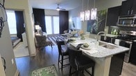 This single-floor space showcases a thoughtfully designed open-concept layout that connects the kitchen and living areas into one cohesive, flowing environment. The kitchen features a curved granite breakfast bar with seating for multiple bar-height chairs upholstered in dark leather with nailhead trim detailing, paired with elegant pendant lighting that hangs above the counter, providing both task lighting and a decorative focal point.

The kitchen is equipped with a modern sink and ample counter space, while the bar area serves as a natural transition into the living space. Rich hardwood flooring runs throughout, grounding the design with warmth and continuity.

The living area is generously proportioned, furnished with a contemporary sofa, an accent chair in a bold animal print, and a geometric patterned area rug that adds visual interest underfoot. A ceiling fan keeps the space comfortable, while large windows dressed in floor-to-ceiling dark drapes allow for adjustable natural light. Wall art featuring lush greenery adds a vibrant, organic contrast to the otherwise sleek, neutral palette. The overall ambiance is modern and sophisticated with carefully curated décor choices.