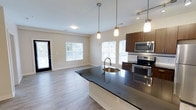 This 2-bedroom, 2-bathroom apartment offers a thoughtfully designed single-floor layout that maximizes both comfort and functionality. The open-concept living and kitchen area serves as the heart of the home, creating a seamless flow between spaces ideal for everyday living and entertaining. The kitchen is outfitted with dark granite countertops, rich dark wood cabinetry, and stainless steel appliances including a range, microwave, and refrigerator. A central island with an undermount sink and pendant lighting above adds both workspace and a stylish focal point to the room. Light wood-style flooring extends throughout the main living area, complementing the neutral, warm-toned walls. Large windows with blinds invite generous amounts of natural light into both the kitchen and the adjoining living area, creating a bright and welcoming ambiance. A glass-panel door provides access to the exterior, further enhancing the connection to the outdoors. The overall design balances modern aesthetics with practical living, making this a well-appointed residence suitable for a variety of lifestyles.