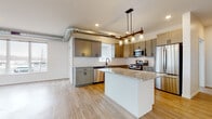 This single-floor unit showcases a contemporary design highlighted by exposed ductwork that adds an industrial character throughout the open living spaces. The kitchen serves as a central hub, featuring gray shaker-style cabinetry with matte black hardware, granite countertops, a white subway tile backsplash, and a full suite of stainless steel appliances including a gas range, built-in microwave, dishwasher, and refrigerator. A central island with a granite surface provides additional workspace and casual seating, while a pendant light fixture with Edison-style bulbs hangs above it, creating a warm focal point. Light wood-look flooring extends through the kitchen and into the spacious living area, which is anchored by a sleek linear electric fireplace and a bank of large windows that invite abundant natural light and exterior views. The bedrooms are carpeted for comfort, each with windows offering outdoor views, and ceiling fans in select rooms. Two bathrooms are finished with clean white cabinetry, white countertops, and shaker-style doors. The primary bathroom features a dual-sink vanity with a large mirror and a walk-in shower, while the secondary bathroom offers a single-sink vanity with ample storage.