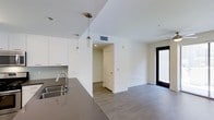 This two-story space showcases a modern, minimalist design characterized by clean lines and a neutral color palette throughout. The open-plan main level integrates the kitchen and living areas into a cohesive, flowing layout ideal for everyday living and entertaining.

The kitchen is outfitted with sleek white cabinetry, a dark quartz or granite countertop, and a stainless steel sink with a contemporary pull-down faucet. Stylish cylindrical pendant lights hang above the counter, providing focused task lighting while adding a decorative touch. A gray tile backsplash complements the overall color scheme.

The adjoining living area continues the modern aesthetic with smooth white walls, recessed lighting, and durable wood-look flooring in a warm gray tone that runs consistently throughout the space. A large floor-to-ceiling black-framed glass door or window floods the area with natural light, enhancing the open, airy feel of the interior.

A doorway leads to an additional room or hallway, offering further functional space within the layout. HVAC venting is discretely integrated into the ceiling, ensuring climate control without disrupting the clean visual design. The overall ambiance is bright, polished, and move-in ready.