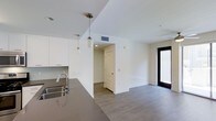 This two-story space showcases a modern, minimalist design characterized by clean lines and a neutral color palette throughout. The open-plan main level integrates the kitchen and living areas into a cohesive, flowing layout ideal for everyday living and entertaining.

The kitchen is outfitted with sleek white cabinetry, a dark quartz or granite countertop, and a stainless steel sink with a contemporary pull-down faucet. Stylish cylindrical pendant lights hang above the counter, providing focused task lighting while adding a decorative touch. A gray tile backsplash complements the overall color scheme.

The adjoining living area continues the modern aesthetic with smooth white walls, recessed lighting, and durable wood-look flooring in a warm gray tone that runs consistently throughout the space. A large floor-to-ceiling black-framed glass door or window floods the area with natural light, enhancing the open, airy feel of the interior.

A doorway leads to an additional room or hallway, offering further functional space within the layout. HVAC venting is discretely integrated into the ceiling, ensuring climate control without disrupting the clean visual design. The overall ambiance is bright, polished, and move-in ready.
