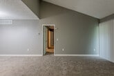 This single-floor residence offers a thoughtfully designed layout with a variety of finishes and architectural details throughout. The living room serves as a central highlight, featuring dramatically vaulted ceilings that lend an open, airy feel to the space. A classic white brick fireplace anchors one corner of the room, adding warmth and character, while vertical blinds covering a sliding door provide access to the outdoors. Plush carpeting runs throughout the living and bedroom areas, contributing to a comfortable atmosphere. Neutral gray tones on the walls create a cohesive, modern palette across the space. The kitchen is well-appointed with white cabinetry, dark granite-style countertops, a stainless steel sink, and a dishwasher, complemented by light wood-look vinyl flooring and a window that brings in natural light. The bathroom features a white vanity with matching dark countertop, a large mirror, and an adjacent open shelving area that offers generous storage. The entry area transitions between carpet and vinyl flooring, and the textured ceilings throughout add a classic residential character. Multiple rooms are accessible via white panel doors, and the overall layout, as seen in the dollhouse view, presents a functional and connected floor plan suited for comfortable everyday living.