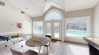 Located at 8104 Webb Rd in Riverdale, GA, this single-room recreational space offers a well-appointed and inviting environment designed for leisure and entertainment. The room is defined by its striking vaulted ceiling, which rises to a dramatic peak and is accentuated by an elegant arched entryway with a fan-lite transom window that floods the space with natural light. Light neutral walls and crisp white trim throughout contribute to a clean, airy atmosphere.

Large windows on multiple sides frame views of an outdoor swimming pool and surrounding greenery, seamlessly connecting the interior to the exterior environment. A set of white French doors with sidelights provides direct access to the pool area, further enhancing the indoor-outdoor flow.

The floor is finished with light-toned square ceramic tiles that reflect natural light and add to the room's bright, open feel. The space is equipped for recreation, featuring a full-sized ping-pong table and a shuffleboard table, making it an ideal gathering spot. A round table with seating occupies a central area, offering a casual space for relaxation or tabletop games. The textured ceiling and dedicated ventilation ensure comfort throughout. This versatile game room presents a functional and welcoming retreat suited for social activities and recreational use.