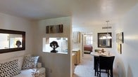 This single-story residence in Euless, Texas, is designed with an open-concept layout that seamlessly connects the primary living spaces. A defining architectural feature is the partial dividing wall between the living room and kitchen, which includes a built-in pass-through window — a functional detail that maintains visual connectivity while subtly defining each zone. The living area is bright and neutral, with light-toned walls and carpeted flooring that carry throughout the space, contributing to a cohesive and understated aesthetic.

The kitchen is equipped with warm wood-toned cabinetry and standard appliances, and flows naturally toward the dining area, which is outfitted with a round table and dark dining chairs beneath a multi-bulb chandelier. A large mirror on the dining room wall enhances the sense of openness and reflects natural light from the nearby window.

Beyond the kitchen, a bedroom is visible, featuring a ceiling fan, shuttered windows that allow for natural light, and warm bedding. The overall ambiance is calm and functional, with neutral tones, modest décor, and a layout that prioritizes flow and practicality throughout the single-floor footprint.