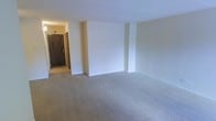 This single-floor space presents a straightforward, functional layout anchored by a spacious open main room. The room features wall-to-wall carpet in a neutral gray tone, providing a clean and understated foundation throughout. The walls are finished in a soft off-white, contributing to a bright and airy atmosphere. Subtle structural columns are integrated into the wall design, adding a modest architectural detail to the otherwise clean-lined interior. The ceiling is flat and smooth, maintaining a consistent height across the main area. A hallway extends from the main room toward the entry, where a solid wood-toned door is visible, framed by light-colored walls that carry the same neutral palette found throughout the space. The overall design is minimal and move-in ready, offering a versatile blank canvas well-suited for a variety of furniture arrangements and uses. The open nature of the main room allows for flexible configuration, making it adaptable to different needs. The connected hallway provides a clear and functional transition between the entry and the primary living area.