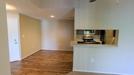 This single-floor residence offers a well-designed open-concept layout that connects the living area and kitchen in a cohesive, functional flow. The living space is finished with rich, warm-toned hardwood flooring that extends throughout, providing a clean and inviting aesthetic. A half-wall partition elegantly defines the kitchen from the living area while maintaining an open, airy feel between the two spaces. The kitchen is equipped with stainless steel appliances, including a gas range and over-the-range microwave, complemented by dark cabinetry and a light-toned countertop that offers a pleasing contrast. Recessed lighting in the ceiling provides focused illumination, while natural light filters in through a window visible in the kitchen area. The living room features neutral, warm-beige walls and transitions to carpeted flooring at the entryway, adding a layer of comfort and texture to the space. The ceiling height lends a sense of openness throughout. Clean white baseboards and trim frame the rooms neatly, contributing to the overall tidy and well-maintained presentation of this practical and comfortable single-level home.