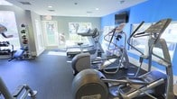 This single-level fitness center offers a comprehensive workout environment designed to accommodate a range of exercise needs. The space is finished with durable rubber flooring in a deep charcoal tone, providing both safety and functionality for high-intensity training. Walls are painted in a calming gray, complemented by a bold blue accent wall that adds visual energy to the environment.

Natural light floods the room through multiple large windows, offering views of surrounding greenery and creating an open, inviting atmosphere. Recessed ceiling lighting supplements the natural light, ensuring the space remains well-illuminated throughout the day.

The cardio section is well-appointed with multiple Precor ellipticals, treadmills, and a stair climber, arranged to maximize floor space and user comfort. A wall-mounted television is positioned for easy viewing during workouts. The free weight area features a full rack of dumbbells alongside a weight bench, and a medicine ball rack provides additional training versatility. A dedicated entrance with a glass-paneled door ensures easy access. A fire safety station is also present, reflecting attention to occupant safety throughout the facility.